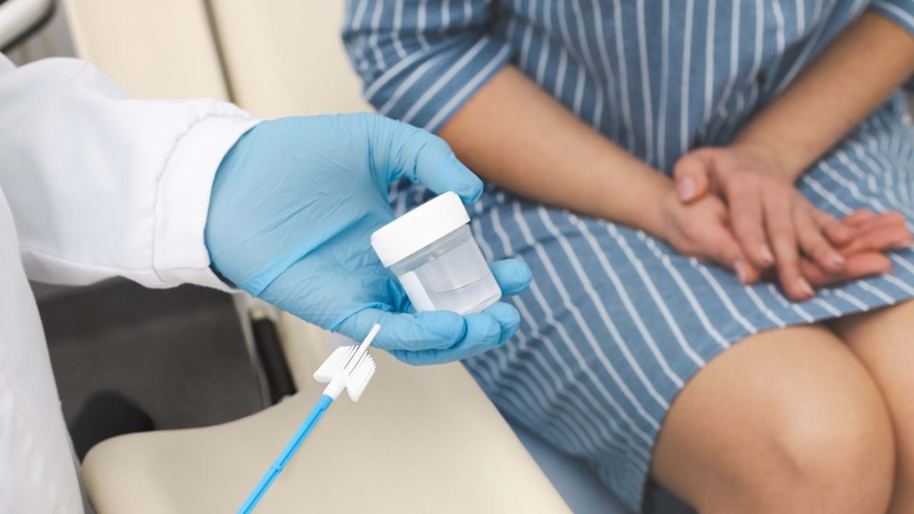 Helsepersonell med blå hansker holder en celleprøvebørste og en beholder for prøvetaking, mens en pasient sitter i bakgrunnen. Bildet illustrerer HPV-screening og livmorhalsprøvetaking.