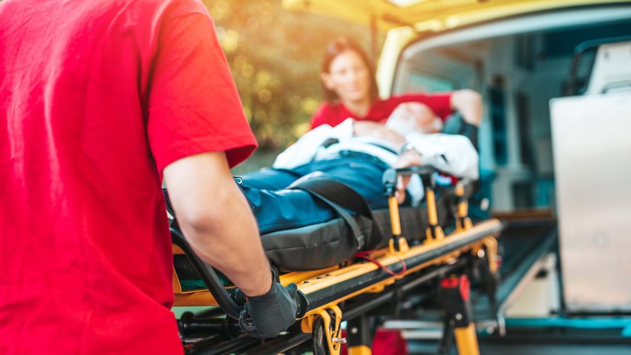 To ambulansearbeidere i røde uniformer laster en pasient på båre inn i en ambulanse. Pasienten er tildekket med et teppe og ser ut til å være bevisstløs.