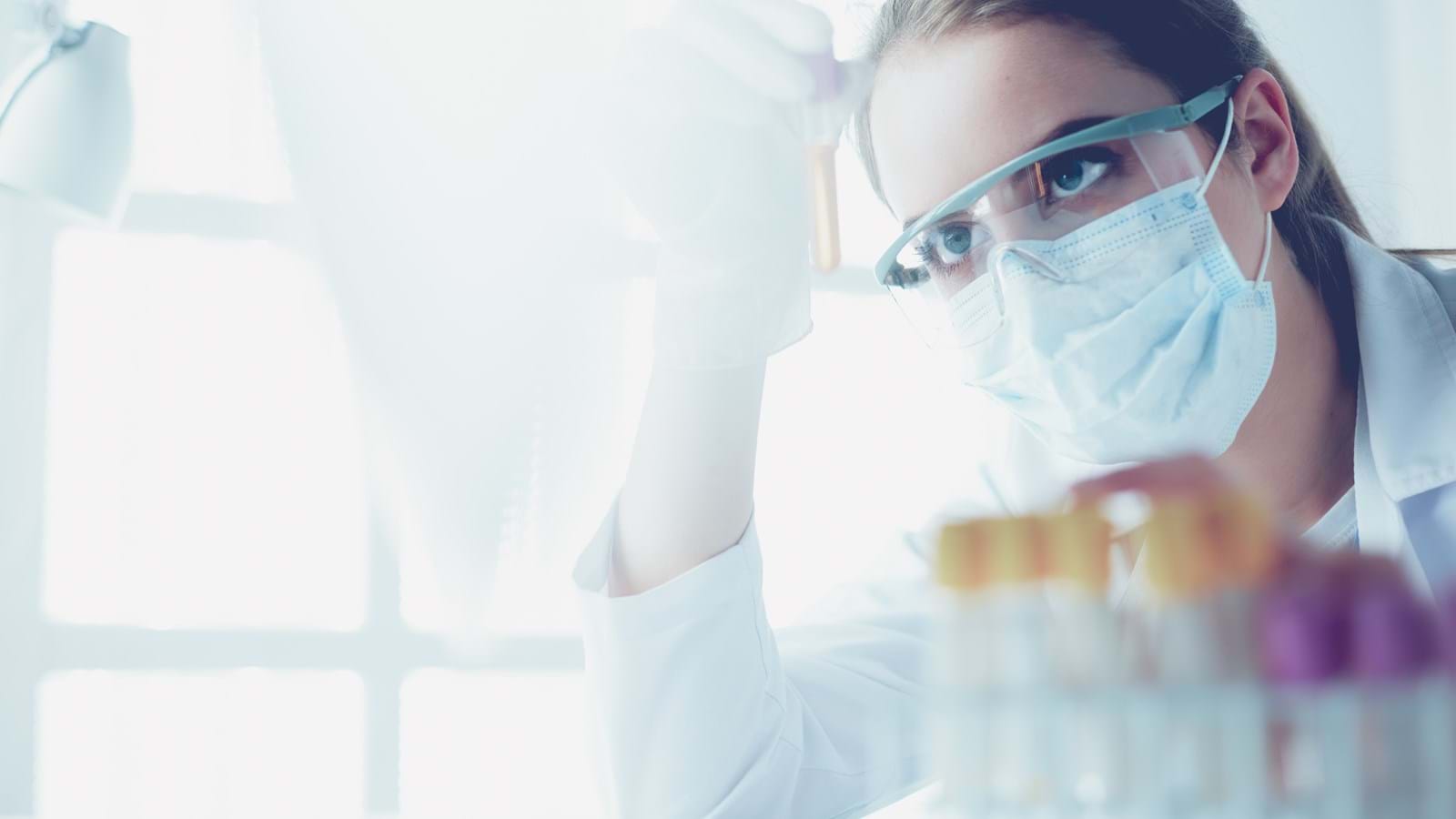 En forsker med vernebriller og ansiktsmaske som undersøker et prøverør i et laboratorium.